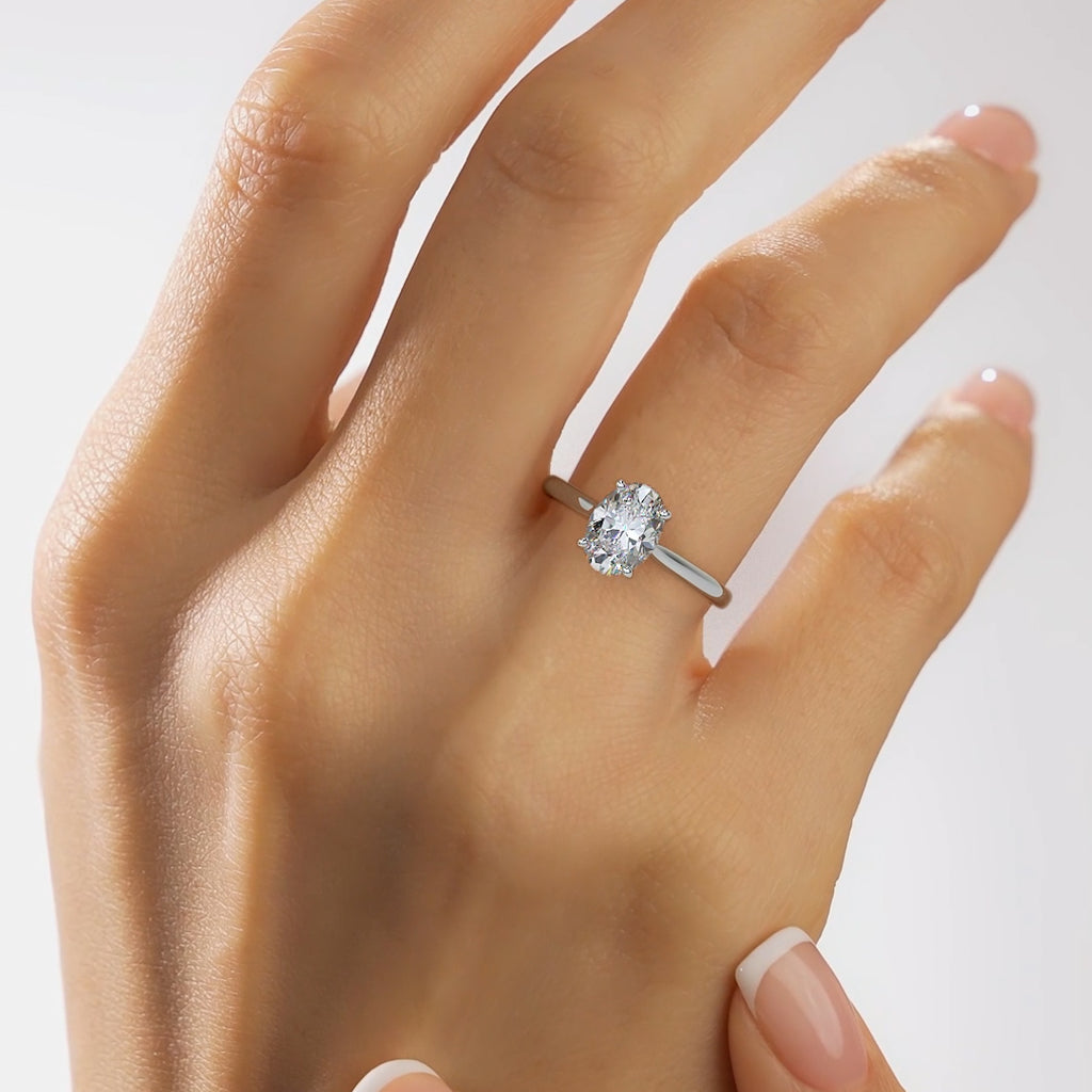 A close-up of a hand wearing a silver engagement ring with a round-cut clear gemstone on a plain light background.