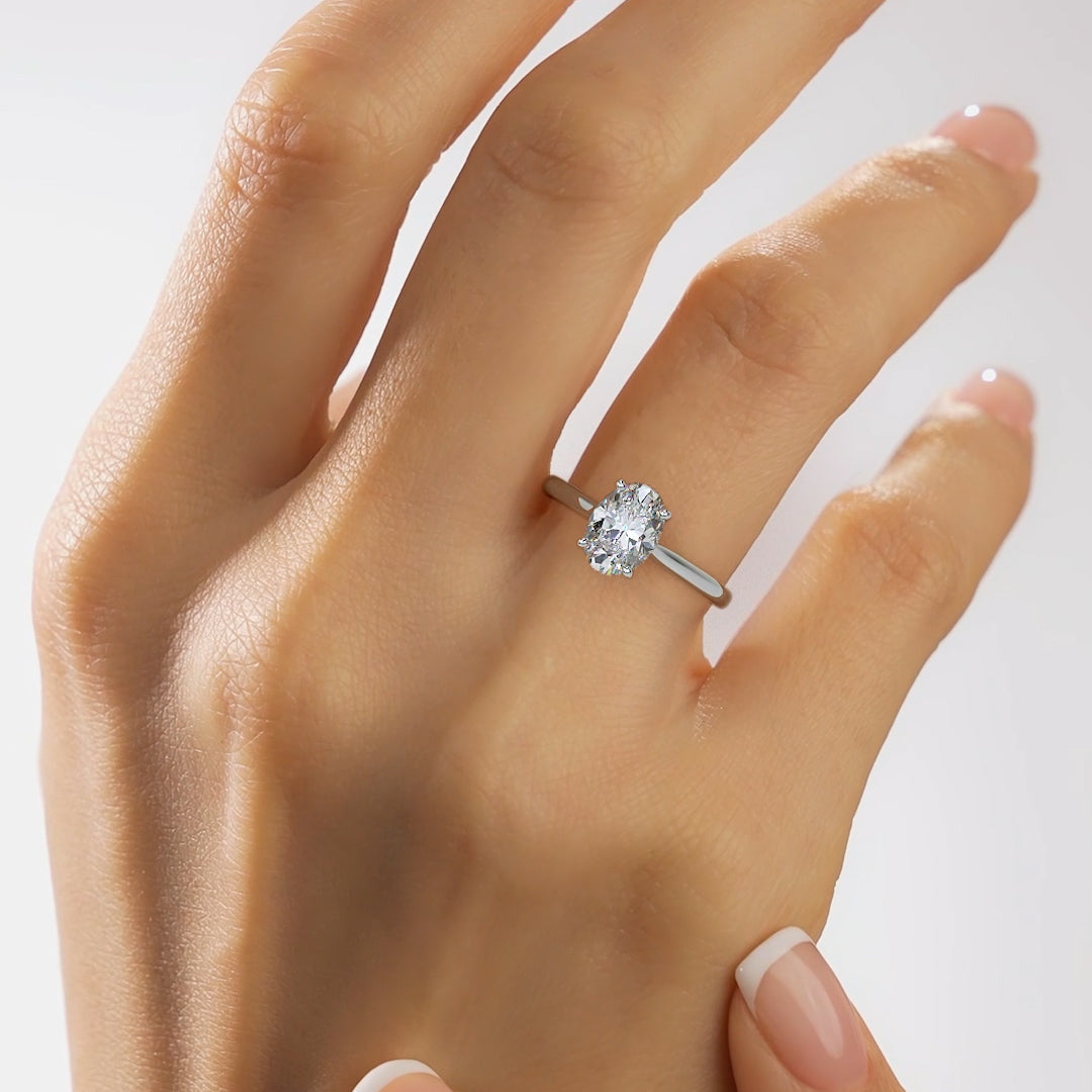 A close-up of a hand wearing a silver engagement ring with a round-cut clear gemstone on a plain light background.