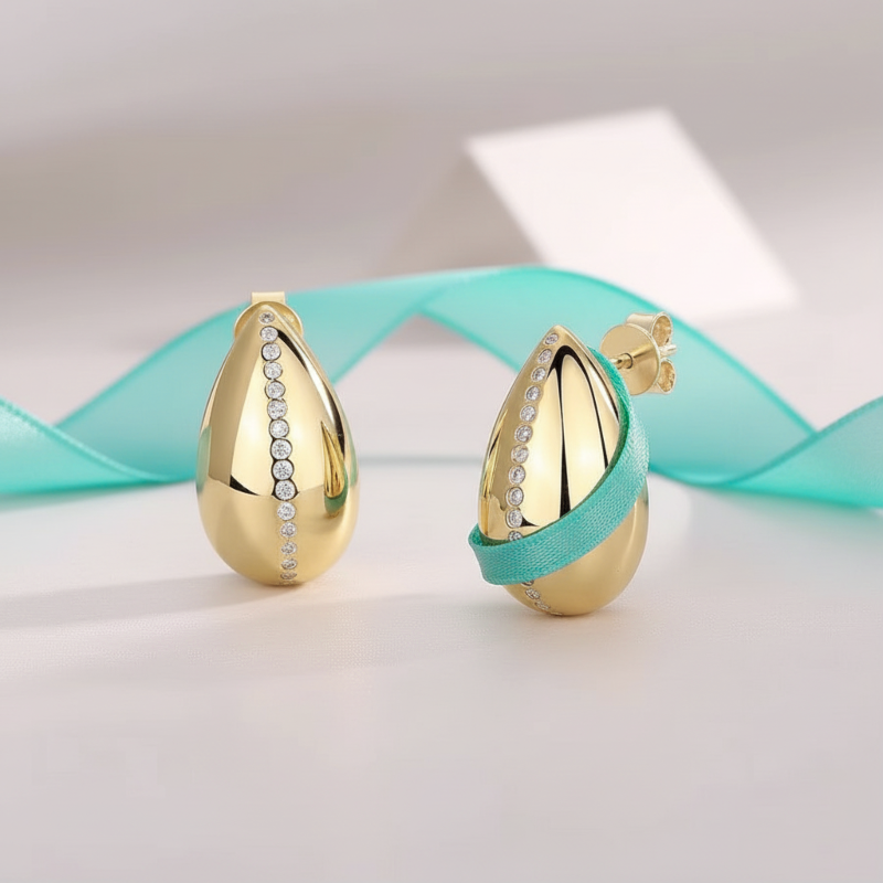 Gold teardrop-shaped earrings with embedded stones on a light background with a turquoise ribbon.
