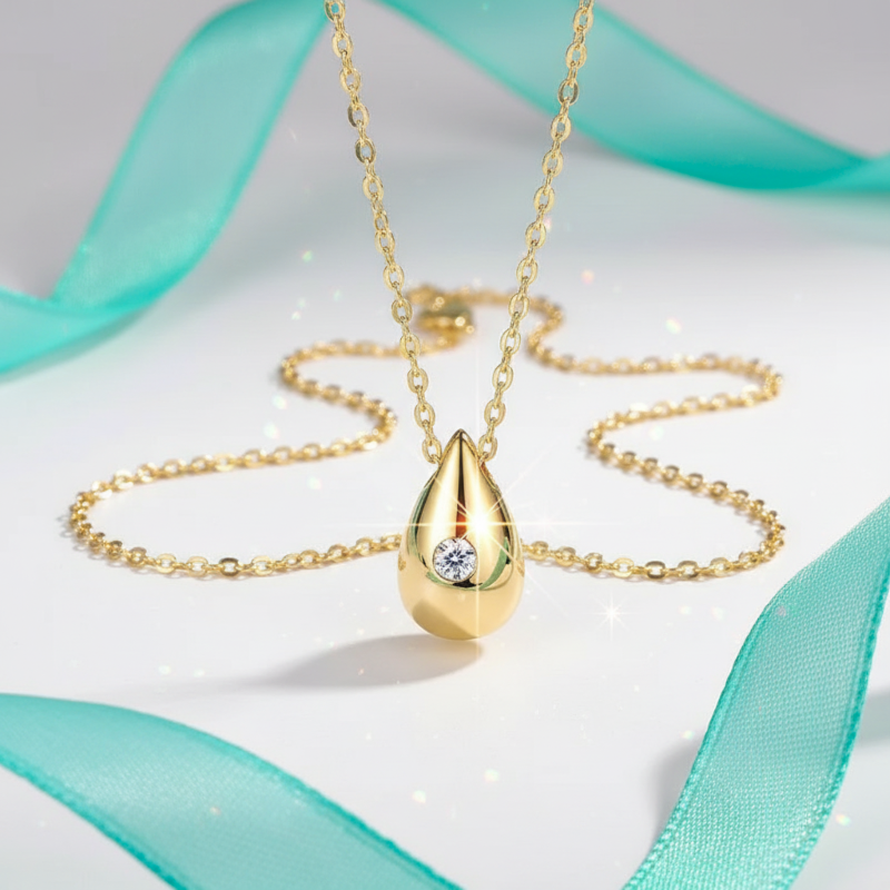 Gold necklace with a teardrop-shaped pendant on a light background with teal ribbons.